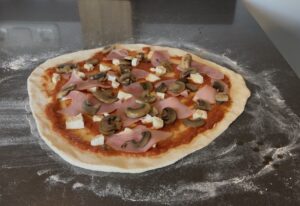 afbeelding van een pizza prosciutto ai funghi belegd voordat deze de oven in gaat